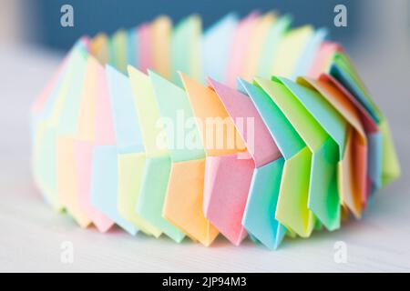 L'anello Origami in fogli di carta colorati collegati giace su un tavolo di legno bianco, foto ravvicinata con messa a fuoco morbida selettiva Foto Stock