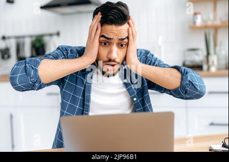 Primo piano di scioccato confuso razza mista giovane uomo, freelancer o dipendente, guardando sorpreso lo schermo del notebook, stressato dal fallimento di un accordo o progetto, leggere notizie inaspettate, in disperazione Foto Stock