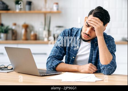 Stanco preoccupato triste arabo o indiano ragazzo, sedersi a una scrivania, guardare stanco e stressato, frustrato guardando i documenti di lavoro, non riuscendo a completare il lavoro sul progetto in tempo, ha bisogno di riposo. Concetto di lavoro eccessivo Foto Stock