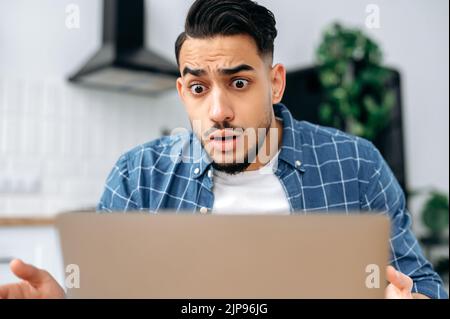 Primo piano di confuso indecolato indiano o arabo, freelance o studente, guardando delusione per lo schermo del notebook, sottolineato dal fallimento di un accordo o progetto, leggere notizie inaspettate, in disperazione Foto Stock