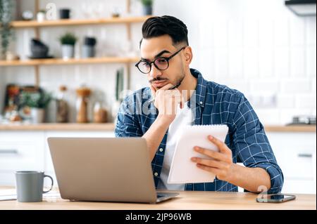 Giovane arabo o indiano moderno e intelligente, che lavora a un progetto mentre si è seduti a casa e si utilizza un notebook, guardando lo schermo con attenzione, pianificando una strategia, analizzando i rischi Foto Stock