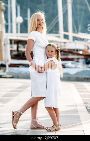 Felice madre millenaria di mezza età e figlia adolescente bionda in abiti bianchi che abbraccia la riva del mare e marina con yacht in un porto turistico Foto Stock