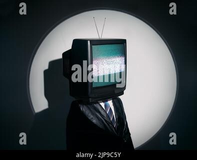 Uomo con la TV invece della testa. Media zombie concetto con televisione addicted maschio in nero cappotto con braccia incrociate in posa in studio sotto i riflettori. Con Foto Stock