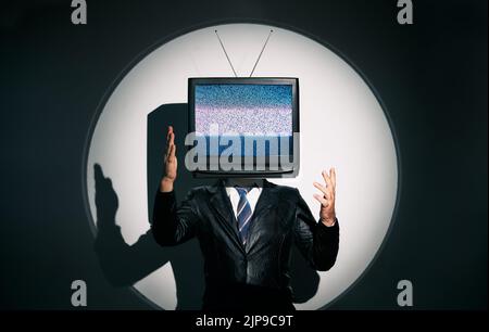 Uomo con la TV invece della testa. Media zombie concetto con televisione addicted maschio in nero cappotto con braccia incrociate in posa in studio sotto i riflettori. Con Foto Stock