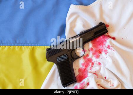 La pistola giace su un abito femminile sanguinoso sulla bandiera dell'Ucraina, guerra in Ucraina, armi, violenza contro gli ucraini 2022 Foto Stock