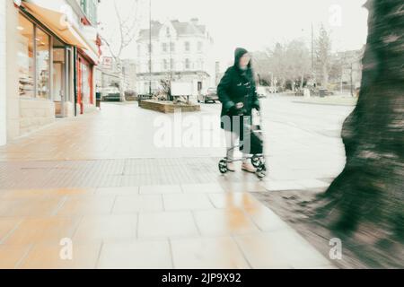 Donna anziana che cammina con una cornice per lo shopping lungo la strada principale nel centro di Ilkley in una giornata invernale piovosa (ICM) West Yorkshire, Inghilterra, Regno Unito Foto Stock