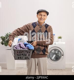 Uomo anziano sorridente che tiene un cesto di biancheria pieno di vestiti all'interno di un bagno Foto Stock