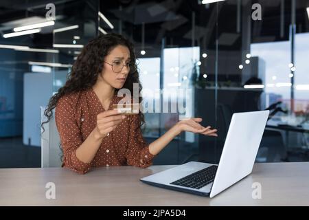 Una donna d'affari sconvolta in un ufficio moderno cercando di fare un acquisto in un negozio online, ha ottenuto un errore e un rifiuto, una donna d'affari che utilizza il laptop e tenendo una carta di credito Foto Stock