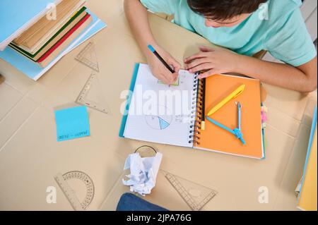 Vista dall'alto di un scolaro che fa un'operazione di geometria matematica sul copybook, studiando da casa in una scuola primaria in linea Foto Stock