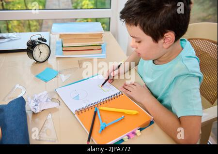 Vista dall'alto di un astuto e concentrato studente caucasico che fa i compiti di geometria, studiando a casa. Concetto di ritorno a scuola Foto Stock