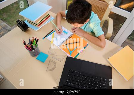 Il ragazzo della scuola risolve esempi di geometria, seduto al tavolo con i rifornimenti della scuola, e il laptop che trasmette video lezione online Foto Stock