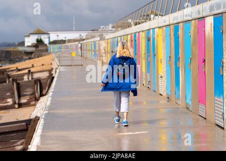 Milford-on-Sea, Hampshire, Regno Unito, 16th agosto 2022, Meteo. Le docce pesanti accompagnano le nuvole scure come le cadute di pioggia necessarie su asciutto, parched l'Inghilterra del sud. Una donna in un impermeabile blu cammina davanti a capanne deserte sulla spiaggia. Credit: Paul Biggins/Alamy Live News Foto Stock