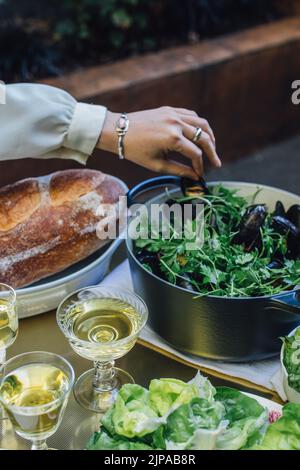 mano di donna che si allunga per prendere il cozze al vapore dal piatto sul tavolo da pranzo all'aperto con vino bianco e pane Foto Stock