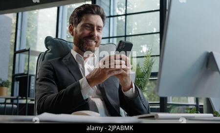 Occupato uomo caucasico barbuto di mezza età 40s uomini d'affari utilizza telefono cellulare app SMS chatting messaggi di testo di lavoro in ufficio scrivania con seduta Foto Stock