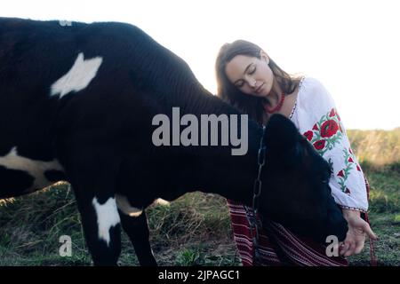 Giovane donna Ucraina si siede e tocca vitello vestito in tradizionale maglia nazionale ricamata e gonna sul pascolo. Vestiti nazionali etnici ucraini Foto Stock