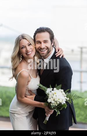 Sposa positiva in abito da sposa che tiene bouquet e abbraccia lo sposo in terrazza Foto Stock