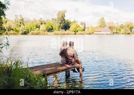Coppia di famiglie anziane che si rilassano sul lago estivo. L'uomo e la donna si godono il paesaggio naturale e si abbraccia seduti sul molo Foto Stock