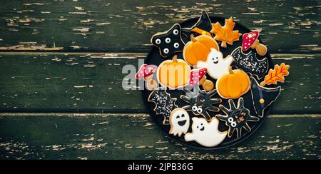 Deliziosi biscotti allo zucchero con glase per Halloween in un piatto - a forma di foglie, zucca, funghi, fantasmi, cappello di strega, ragno. Vista dall'alto. Sfondo verde scuro con spazio di copia Foto Stock