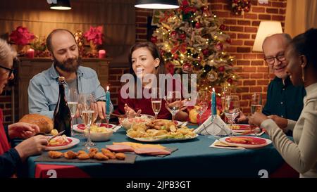 Moglie felice che parla con gli ospiti del cibo mentre si siede insieme al tavolo della cena di Natale. Festa gente allegra godendo tradizionale pasto di festa di inverno mentre festeggia la festa. Foto Stock