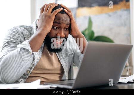 Irritato arrabbiato stressato afroamericano uomo, sedersi alla scrivania in ufficio, utilizzando il portatile per il lavoro online, infastidito con il cattivo risultato del suo progetto, non riuscito l'accordo, esperienze di emozioni negative, stress Foto Stock