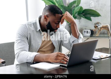 Triste esausto afro americano giovane uomo, dipendente aziendale, seduto alla scrivania, sembra stanco e stressato, l'esperienza di mal di testa, emicrania, ha bisogno di riposo, ha chiuso gli occhi. Lavoro in eccesso, scadenza Foto Stock