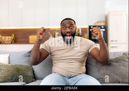 Eccitato gioioso attraente giovane afro-americano, seduto sul divano in soggiorno, eccitato per il successo con le braccia alzate, celebrando la vittoria sorridendo felicemente, urlando. Concetto vincente Foto Stock