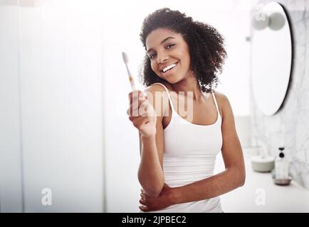 Il tuo sorriso è la curva che fissa tutto dritto. Ritratto di una giovane donna attraente spazzolando i denti nel bagno a casa. Foto Stock