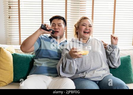 Giovane coppia asiatica chubby che canta e guarda la tv sul divano. L'uomo e la donna si divertono insieme a casa. Le persone ridono e sorridono insieme Foto Stock