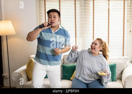 Giovane coppia asiatica chubby che canta e guarda la tv sul divano. L'uomo e la donna si divertono insieme a casa. Le persone ridono e sorridono insieme Foto Stock