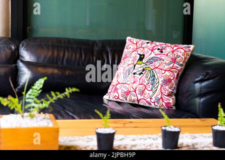 Il cuscino rosso con motivi Peacock si trova su un divano nero. Davanti a lui c'era una piccola pianta in vaso su un tavolo di legno marrone. Foto Stock