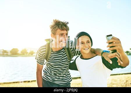 Venite a prendere un selfie con me, due giovani fratelli che scattano selfie all'esterno con una laguna sullo sfondo. Foto Stock