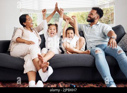 Genitori felici e bambini eccitati festeggiano sul divano mentre guardano la tv. Divertimento in famiglia, relax a casa e legame. Madre e padre alto cinque Foto Stock