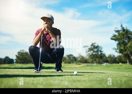 Sul fairway. Un giovane golfer maschio messo a fuoco che guarda una sfera di golf mentre è seduto sull'erba fuori durante il giorno. Foto Stock