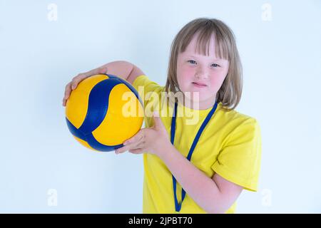 Ritratto di una ragazza carina con occhi blu e capelli biondi con sindrome di Down in una T-shirt gialla tiene una palla da pallavolo giallo-blu nelle sue mani isolate Foto Stock