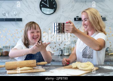 Primo piano di una ragazza con sindrome di Down e la sua affascinante madre, la cottura torte pasta in cucina della loro casa e sorridente allegramente. Il concetto di m Foto Stock