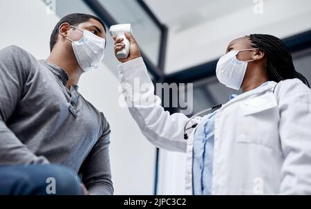 Medico di sesso femminile che consulta un paziente di sesso maschile, prendendo la sua temperatura con termometro a infrarossi in sala o clinica ospedaliera. Uomo che indossa la maschera e. Foto Stock