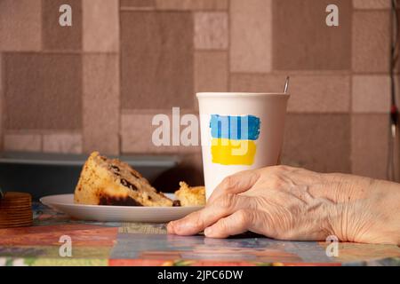 La bandiera giallo-blu dell'Ucraina è dipinta su una tazza bianca con caffè accanto alla mano di una donna e una torta nella cucina di una casa in Ucraina, fermare il Foto Stock