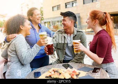 Donna felice ridendo e abbracciando diversi amici con la birra seduto al tavolo da pub con spuntini e smartphone sulla strada della città Foto Stock