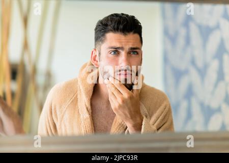 Giovane maschio in caldo accappatoio che tocca la barba e guardare in specchio durante la routine di cura della pelle al mattino nel salone termale Foto Stock