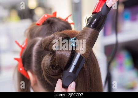 Le pinze calde per primo piano arricciano una ciocca di capelli sulla testa di una donna. Il parrucchiere crea un'acconciatura per una giovane donna. Barbiere, concetto di business. Salone di bellezza, cura dei capelli. Foto Stock