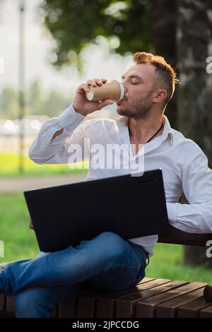 Uomo felice lavora nel parco con un computer portatile, bevande caffè. Un giovane su uno sfondo di alberi verdi, una calda giornata estiva di sole. Luce calda e soffusa, primo piano. Foto Stock