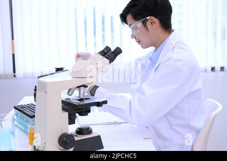 Giovane scienziato maschile asiatico concentrato o specialista che lavora nel suo laboratorio d'ufficio, lavorando sul microscopio e sulla carta di ricerca. Foto Stock