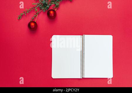 Quaderno a spirale aperto vicino al ramo dell'albero di Natale con decorazioni su sfondo rosso alla moda. Copia testo spazio, elenco cose da fare, sogni. Pianificazione 2023, scrittura nuovo anno wishlist concetto. Stile minimalista. Foto Stock