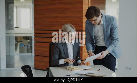 Due colleghi di lavoro discutono di report in un ufficio moderno al chiuso. Donna d'affari seduta al tavolo parlando il suo compagno maschio Foto Stock