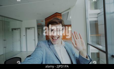 POV di un giovane uomo d'affari in tuta che ha video chat online utilizzando la fotocamera dello smartphone e parlando con i suoi colleghi in un ufficio moderno al coperto Foto Stock