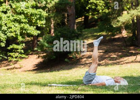 un uomo anziano in abbigliamento sportivo che si stalla a spalla sul tappetino per il fitness nel parco verde Foto Stock