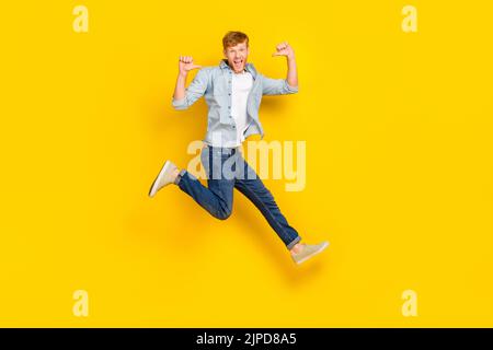 Foto di dimensioni complete di fortunato bel ragazzo rosso capelli indossare camicia blu jeans sneakers vendita impressionato salto isolato su sfondo giallo colore Foto Stock