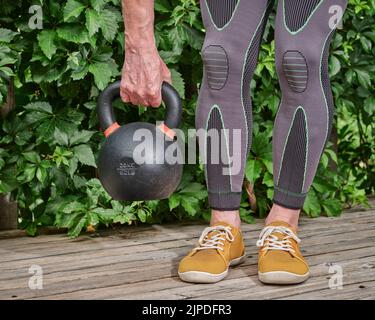 maschio indossare ginocchiere di compressione e calzini bretelle è l'esercizio con un pesante kettlebell di ferro su un ponte di legno cortile Foto Stock