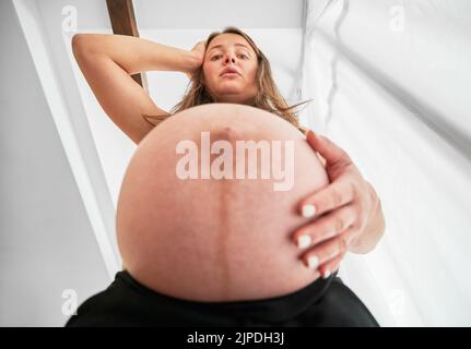 Vista dal basso della donna incinta che ha sorpreso per le dimensioni del suo ventre. Donna che mette una mano sul ventre incinta e l'altra sulla testa e che guarda giù con sgomento. Foto Stock