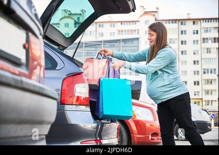 Vista laterale di una donna incinta con ventre che si gonfio borse della spesa nel tronco dell'automobile godendo la maternità e la paternità in modalità di attesa, donna incinta che si prepara per la nascita del bambino durante il Venerdì Nero Foto Stock